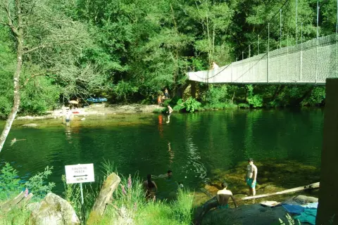 Puente colgante sobre río verde con gente bañándose y disfrutando del paisaje.