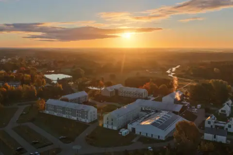 Zachód słońca nad nowoczesnym kompleksem budynków, przypominającym spokojny folwark lochów, otoczonym jesienną przyrodą i rzeką.
