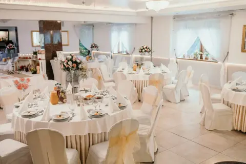 Elegancka sala weselna przygotowana na przyjęcie dla 100 osób. Stoły nakryte białymi obrusami, ozdobione kwiatami i zastawą. Krzesła w pokrowcach z kokardami.
