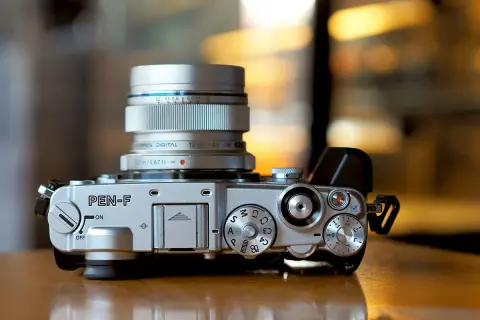 Silver Olympus PEN-F camera with a silver Olympus Digital 12mm 1:2.0 lens attached. The camera body features various dials and buttons, ready for capturing stunning images with its Olympus PEN F lenses.