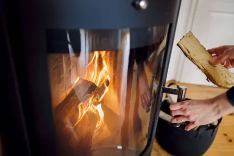 Eine Hand legt Holzscheite nach, um das Feuer im Kamin zu verbrennen. Die Flammen tanzen im Inneren des Ofens.