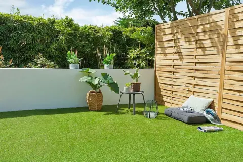 Un jardín moderno con césped artificial verde vibrante, rodeado de plantas y una valla de madera. Un rincón acogedor con cojines invita al descanso.