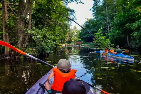 Rodzina płynie kajakiem po malowniczym Pojezierzu Brodnickim, otoczona bujną zielenią.