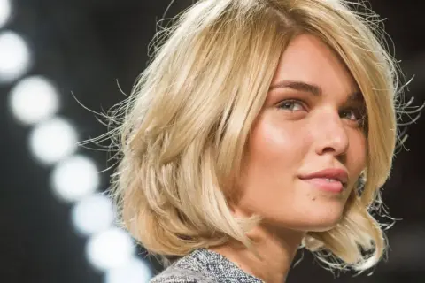 Blonde Frau mit einem stufigen Bob und dicken Haaren, die im Scheinwerferlicht glänzen.