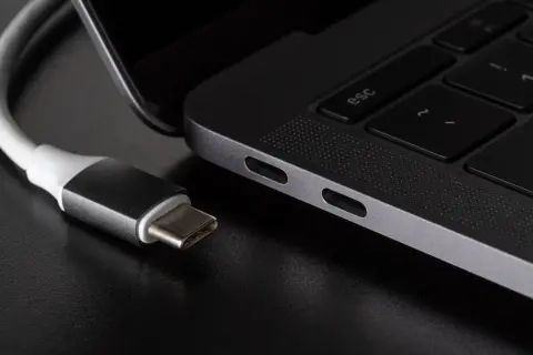 Kabel USB C zbliża się do portu laptopa. Widać jego metalową wtyczkę i biały kabel.