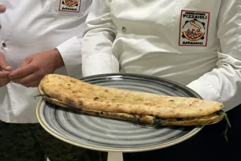 Una paposcia pugliese, ripiena e dorata, servita su un piatto grigio. Indossano giacche con stemmi dell'Associazione Pizzaioli Garganici.