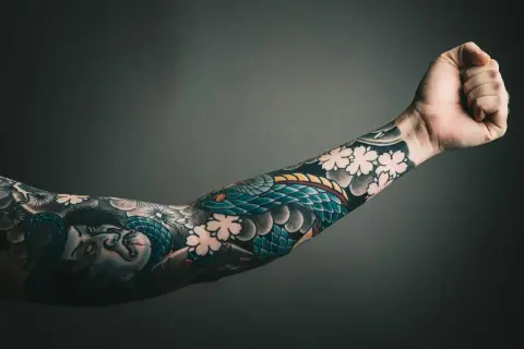 Ein Arm mit einem detailreichen, **heavy blackwork tattoo** von einer Schlange und einer Figur, umgeben von Kirschblüten.