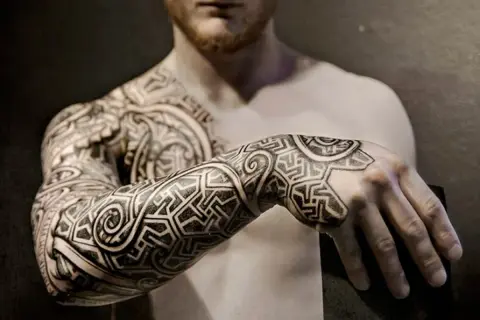 Mann mit beeindruckenden Sleeve Tattoo Ideen, die sich über Arm und Schulter erstrecken. Geometrische Muster und geschwungene Linien.