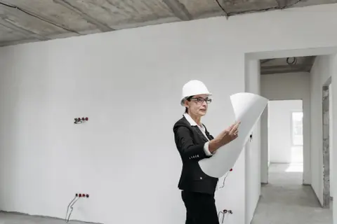 Kobieta w kasku i okularach analizuje plany budowy w niedokończonym wnętrzu.