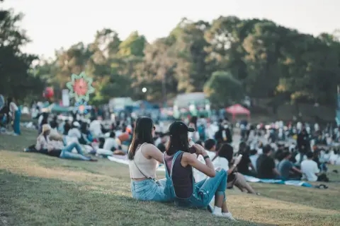 Dwie dziewczyny siedzą na trawie, ciesząc się atmosferą festiwalu. W tle widać tłum ludzi i kolorowe dekoracje, zapowiadające wspaniałe igo koncerty.