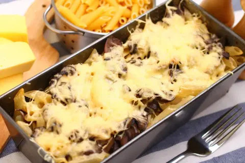 Pyszna zapiekanka z pieczarkami i makaronem penne, zapieczona pod złotym serem. Obok surowy makaron i kawałki sera.