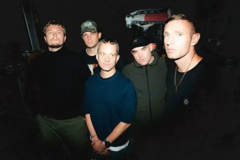 Five men stand in a dimly lit room, ready to perform. One holds a high vis setlist, hinting at the night's energetic show.