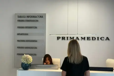 Recepcja kliniki PRIMAMEDICA z tablicą informacyjną i pracownicą.