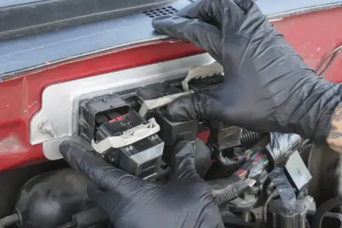 Manos enguantadas trabajan en la caja de fusibles, conectando un componente que podría ser el PCM, el cerebro del vehículo.