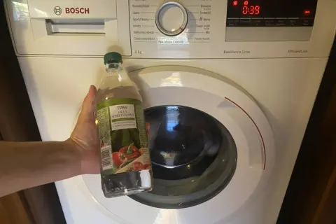 Ręka trzyma butelkę octu spirytusowego obok pralki Bosch, przygotowując się do czyszczenia pralki octem.