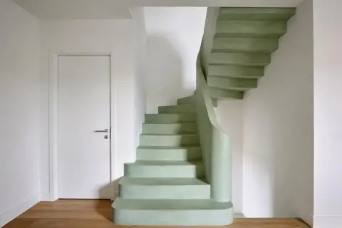 Escaleras curvas de microcemento en tono verde menta, con una puerta blanca y suelo de madera.