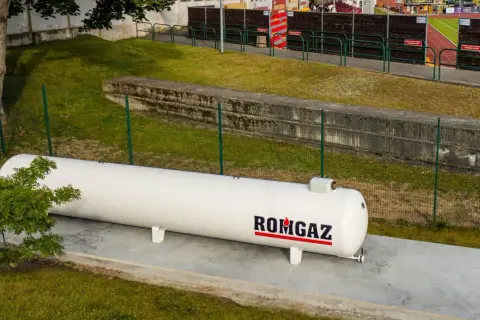 Biały zbiornik gazu ROMGAZ stoi na betonowej podstawie. Widać ogrodzenie i trawę, a w tle trybuny stadionu. Odległość gazociągu od ogrodzenia jest widoczna.