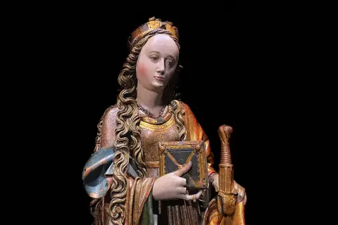 Drewniana figura świętej z koroną, księgą i mieczem na czarnym tle.