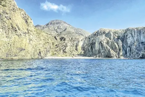 Aguas azules bañan la costa rocosa de una cala en Almería. Montañas escarpadas y una pequeña playa de arena componen el paisaje.