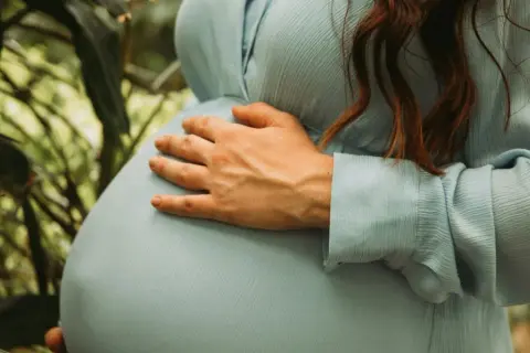 Gravid i vecka 5, en kvinna med ljusblå klänning håller om sin mage bland gröna blad.