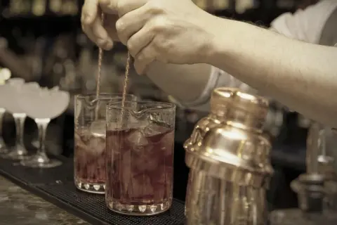 Barkeeper mixt einen roten Bier Cocktail mit Eis in einem Glas. Daneben steht ein goldener Shaker.
