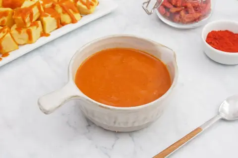 Un cuenco de cremosa salsa brava, lista para acompañar unas patatas. En el fondo, pimentón y guindillas secas.
