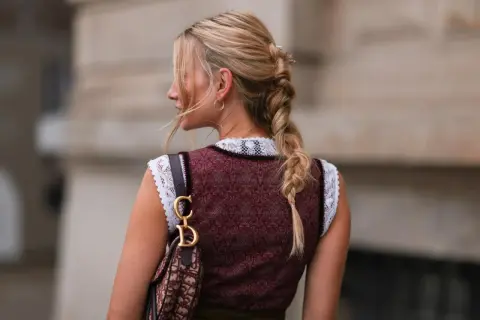 Blonde Zopf-Frisur mit Blumenakzenten, perfekt für Oktoberfest Frisuren. Trägt Dirndl und eine Designer-Tasche.