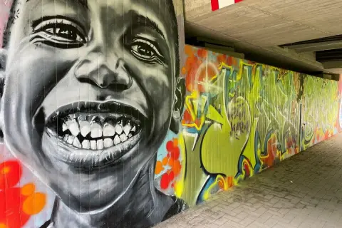 Graffiti-Porträt eines lachenden Kindes neben einer bunten Wand mit Schriftzügen.