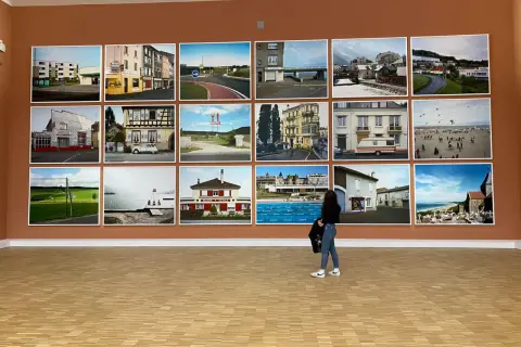 Mostra di fotografie di Raymond Depardon: paesaggi urbani, rurali e marittimi, con una donna che osserva le opere.