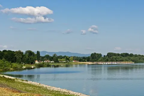 Spokojne jezioro otmuchowskie z piaszczystą plażą, otoczone zielenią. W oddali widać góry i wiatraki.