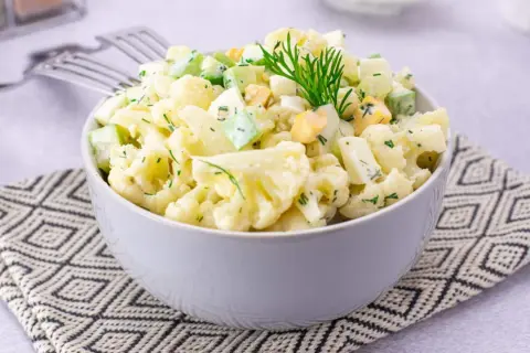 Sałatka z kalafiorem, jajkiem i ogórkiem, udekorowana koperkiem, podana w misce na wzorzystej serwetce.
