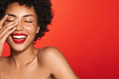 Une femme rit, la main sur le visage, avec un rouge à lèvres éclatant. Quelle couleur de rouge à lèvres !