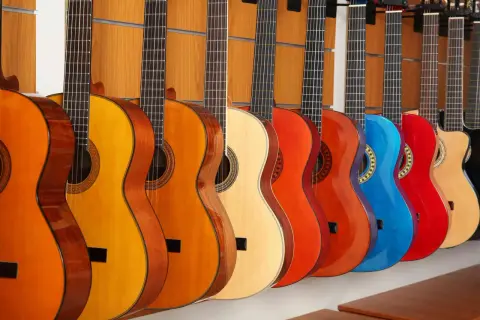 Rząd kolorowych instrumentów akustycznych, w tym gitar w odcieniach pomarańczu, żółci, czerwieni i niebieskiego, wisi na drewnianej ścianie.