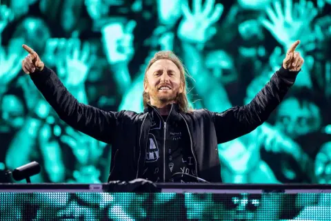 David Guetta na jednym ze swoich koncertów, z rękami uniesionymi w geście radości. Tłum na tle ekranów LED.