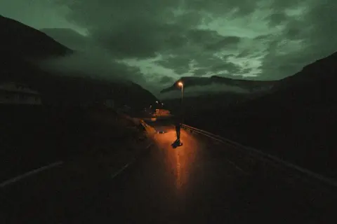 Une silhouette solitaire sur une route de montagne, éclairée par un lampadaire. Le ciel est sombre et nuageux, donnant à cette **grain photo en ligne** une atmosphère mystérieuse.