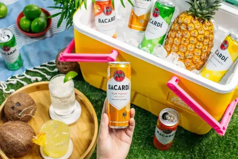 Letnia piknikowa atmosfera z drinkami Bacardi. W żółtej lodówce schłodzone puszki rumowych koktajli i ananas. Na trawie dwa drinki, kokosy i limonki.
