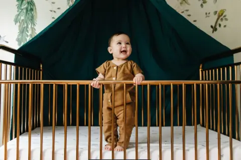 A happy baby stands in a golden crib, holding the bars. What do you need for a crib? A safe space, a soft mattress, and a smiling baby!