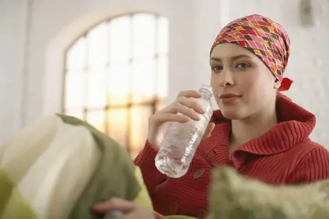 Kobieta w chustce na głowie pije wodę podczas chemioterapii na raka.