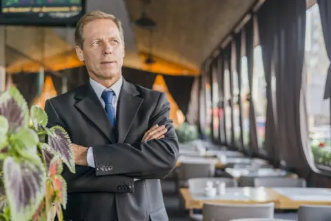 Uomo d'affari con braccia conserte, un chief people officer che guarda lontano, con piante verdi in primo piano e un ristorante sullo sfondo.