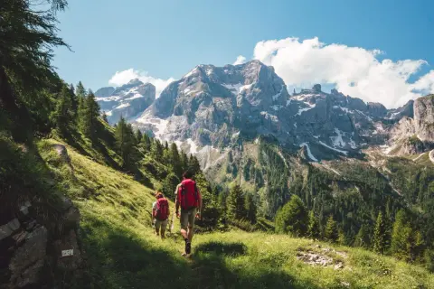 Dos personas disfrutan del senderismo y trekking por un sendero verde con imponentes montañas nevadas de fondo.