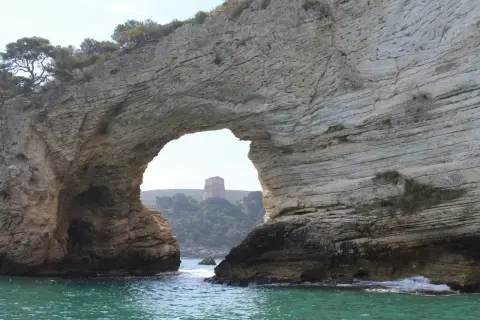 Arco naturale di roccia che incornicia una torre sulla collina, vista dalla baia San Felice Vieste. Acque turchesi e vegetazione mediterranea.