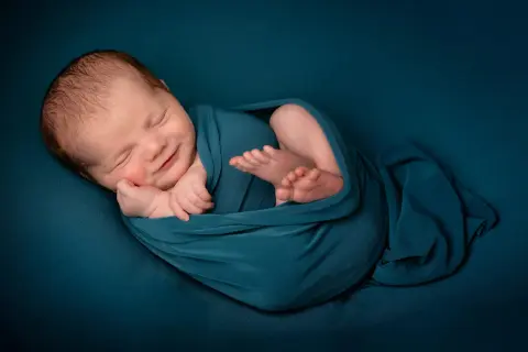 Un bébé endormi, emmailloté dans un tissu bleu canard, sourit paisiblement. Une belle image pour célébrer une naissance, un moment de joie et de gratitude, que dire pour une naissance islam.