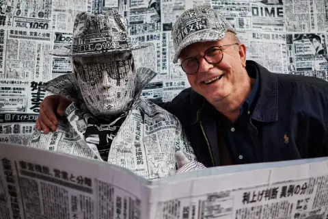 Tomasz Tomaszewski w kapeluszu i płaszczu z gazet. Obok uśmiechnięty mężczyzna w okularach.