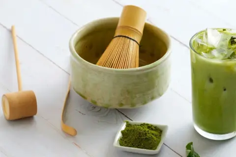Przygotuj pyszny matcha latte! Na stole leży bambusowa miotełka, łyżeczka, miseczka z zielonym proszkiem matcha i szklanka orzeźwiającego napoju.