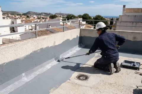 Persona impermeabiliza terraza con rodillo, aplicando pintura gris sobre malla de refuerzo.