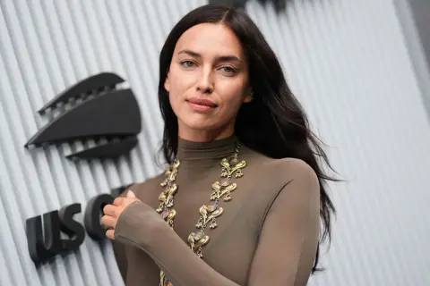Irina Shayk, con un look audace e una collana d'oro, posa davanti a un logo.