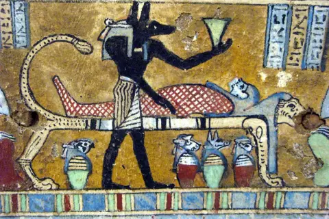 Anubis, bóg z głową szakala, stoi przy katafalku z mumią. W ręku trzyma naczynie, a obok stoją urny z głowami zwierząt.