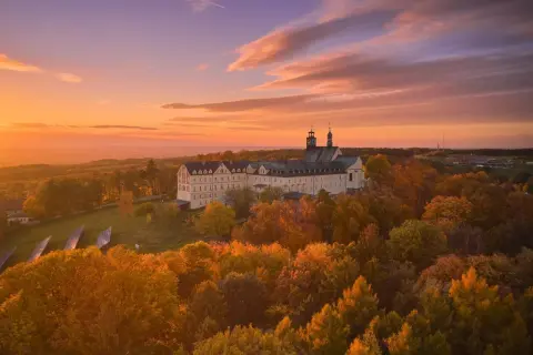 Góra Świętej Anny w jesiennej szacie, otoczona złotymi liśćmi, z majestatycznym klasztorem w tle. Zachód słońca maluje niebo odcieniami pomarańczy i fioletu.