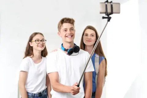 Trzech przyjaciół robi sobie selfie z kijkiem. Chłopak z przodu uśmiecha się szeroko, dziewczyna w okularach po lewej też, a ta z tyłu patrzy w obiektyw. To prosty sposób, jak zrobić ładne zdjęcie grupowe.