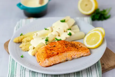 Ein saftiges Stück Lachs liegt neben weißem Spargel mit Sauce Hollandaise auf einem Teller. Garniert mit Petersilie und Zitronenscheiben.